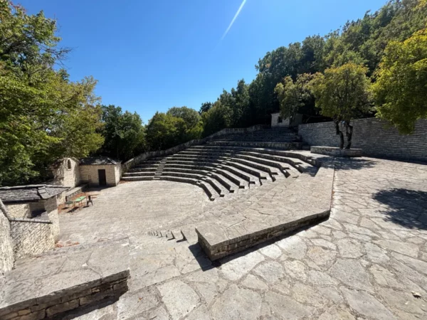 Ancient stone amphitheater surrounded by lush trees in Monodendri, Zagorochoria, Ioannina, Greece. Clear blue sky above.
