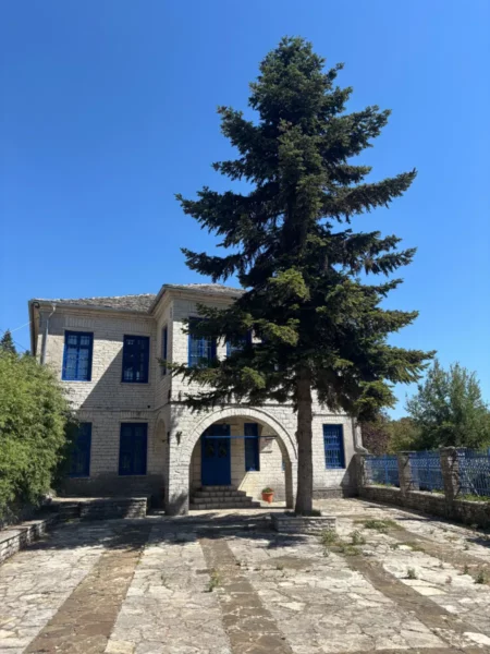 Stone-built school building in Monodendri, Zagorochoria, with blue-painted doors and windows, a paved courtyard, and a tall cypress tree rising in front under a clear summer sky.