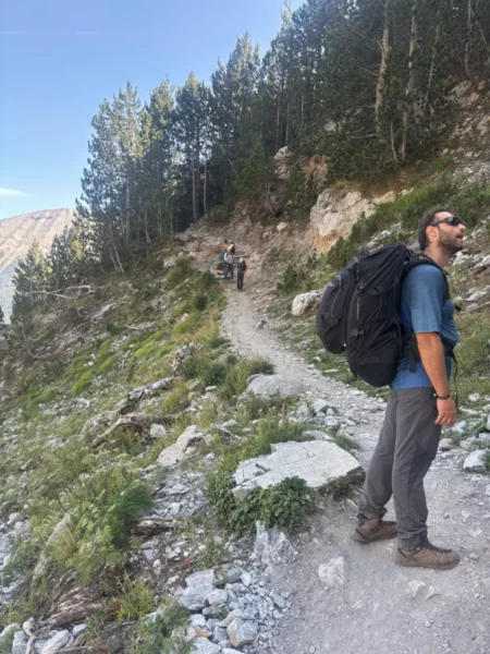 Hiker pauses on a rocky trail surrounded by lush greenery and towering pine trees near Mount Olympus, Pieria, Greece.