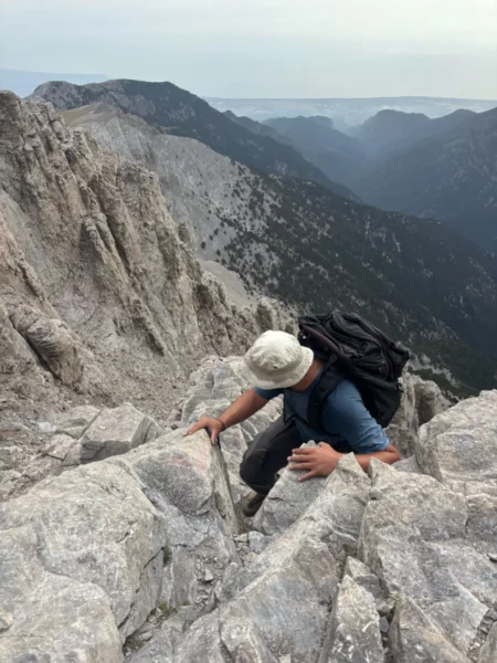 Hiker scrambling along the Kakoskala route on Mount Olympus, using hands on steep limestone terrain between Skala and Mytikas.