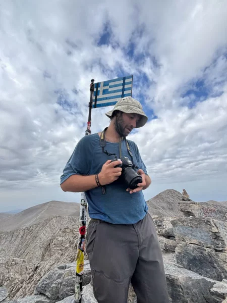 A traveler stands on Mount Olympus in Pieria, Greece, holding a camera, with a Greek flag waving above him against a cloudy sky.