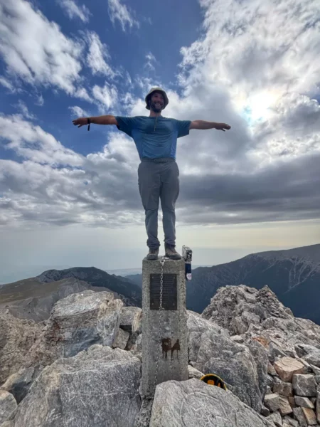 A person stands triumphantly on a stone pillar atop Mount Olympus, surrounded by rocky peaks and cloudy skies in Pieria, Greece.