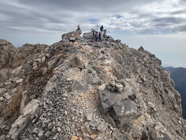 Hikers standing on the narrow summit ridge of Mytikas, the highest peak of Mount Olympus, with jagged limestone and steep drops on both sides.