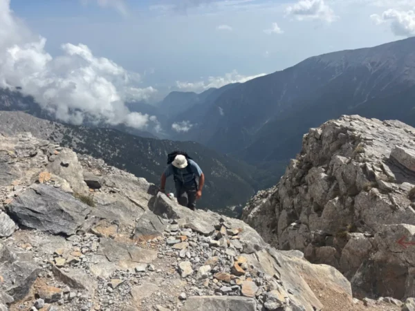 A hiker ascends rocky terrain on Mount Olympus, surrounded by dramatic cliffs and clouds in Pieria, Greece.