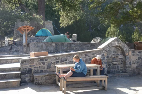 Travelers relax at a stone seating area near tents, surrounded by greenery at Mount Olympus, Pieria, Greece.