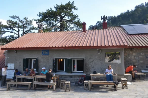 Spilios Agapitos refuge on Mount Olympus, with hikers resting at outdoor tables in front of the stone building just above the treeline.