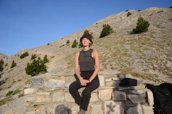 A woman sits on a stone wall against the scenic backdrop of Mount Olympus in Pieria, Greece, surrounded by rugged terrain.