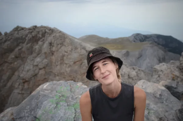 Smiling woman wearing a black hat sits on rocky terrain with Mount Olympus in the background, Greece.