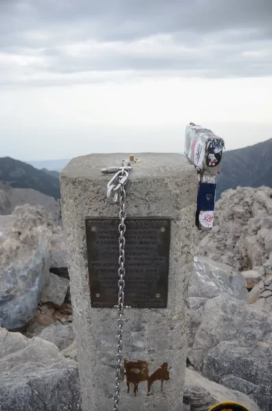 Summit marker at Mount Olympus, Pieria, Greece, featuring a plaque, chains, and surrounding rocky terrain under a cloudy sky.