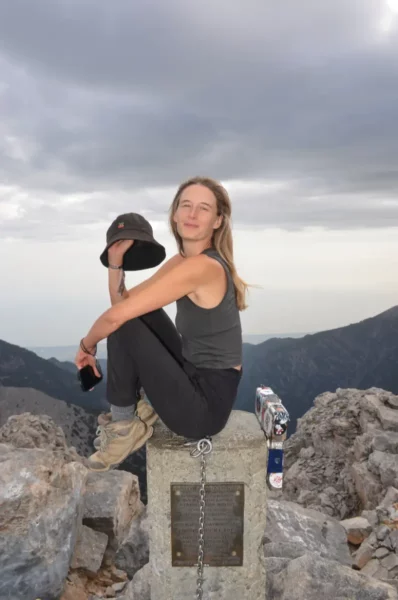 A young woman sits on a stone marker at Mount Olympus, Pieria, Greece, with a mountainous landscape and cloudy sky in the background.