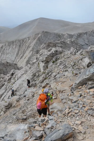 Hikers ascend rocky terrain on Mount Olympus, surrounded by rugged cliffs and a muted, overcast sky in Pieria, Greece.