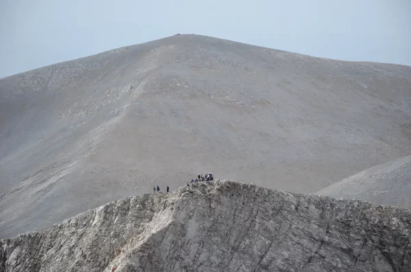 Dramatic view of Mount Olympus in Pieria, Greece, showcasing its rocky summit and distant hikers against a gray sky.
