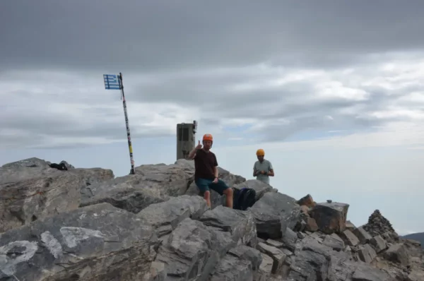 Climbers resting on rocky terrain at Mount Olympus, Greece, with a flag and cloudy skies in the background.
