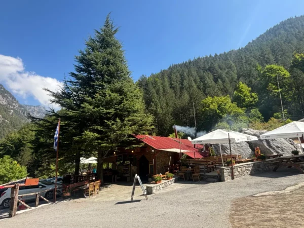 Prionia trailhead on Mount Olympus, with a mountain tavern, outdoor seating, and dense fir forest on the lower slopes.