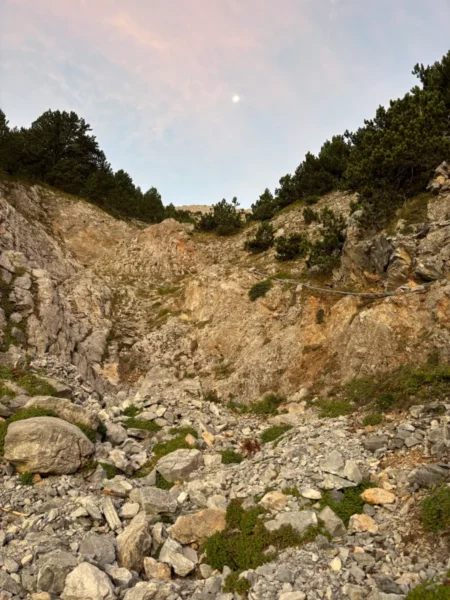 Rocky terrain with scattered boulders and green vegetation, under a pastel sky at Mount Olympus, Pieria, Greece.