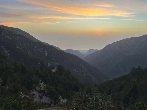 Mount Olympus in Pieria, Greece, showcases a stunning sunset over rugged valleys and distant hills, bathed in soft golden hues.