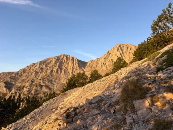 Sunlight illuminates the rugged peaks of Mount Olympus in Pieria, Greece, surrounded by greenery and rocky terrain.