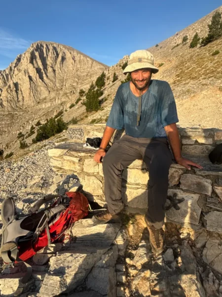 Hiker resting on a stone structure with Mount Olympus in the background, showcasing the rugged terrain of Pieria, Greece.