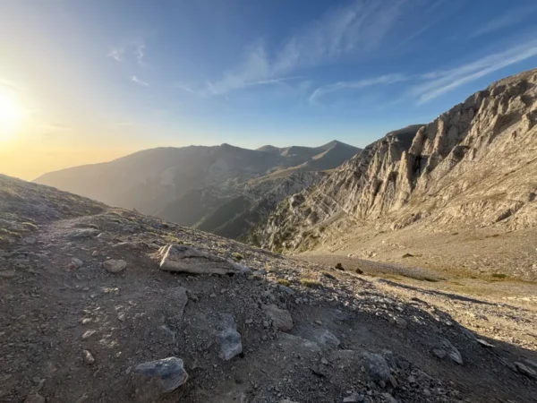 Sun rising over the Aegean Sea as seen from the upper slopes of Mount Olympus, with rocky alpine terrain in the foreground.