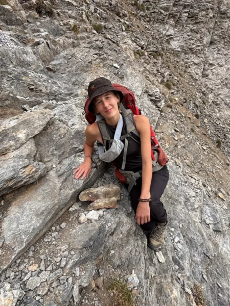 A hiker rests on rocky terrain near Mount Olympus in Pieria, Greece, showcasing a rugged mountain landscape.