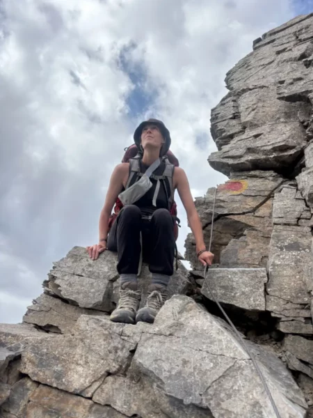 Adventurer perched on rocky summit of Mount Olympus, surrounded by dramatic clouds and rugged stone, near Pieria, Greece.