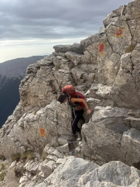 Climber navigating rocky terrain on Mount Olympus in Pieria, Greece, surrounded by rugged cliffs and cloudy skies.