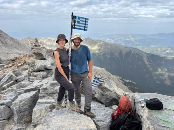 Travelers proudly hold a Greek flag atop Mount Olympus in Pieria, Greece, surrounded by rocky terrain and stunning mountain views.