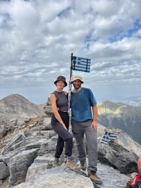 Two hikers standing on the summit of Mytikas, the highest peak of Mount Olympus, next to a Greek flag, with rocky alpine terrain and distant peaks under a cloudy sky.