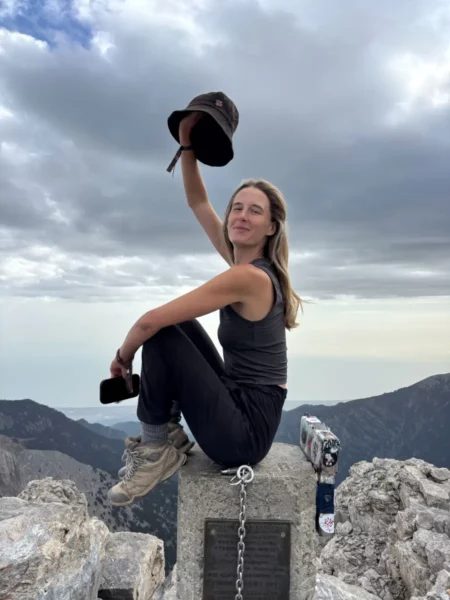 A traveler sits on a mountain summit, raising a hat with a dramatic sky above and scenic views of Mount Olympus, Pieria, Greece.