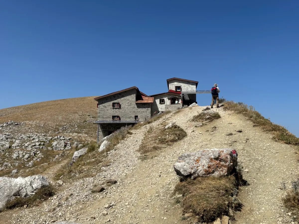 Hiker approaching the Astraka mountain refuge on a rocky ridge below Gamila Peak, Mount Tymfi, with barren alpine terrain and clear blue sky.