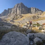 Hiker ascending a rocky alpine trail on Mount Tymfi in the Pindus Mountains, with Gamila Peak and limestone cliffs rising under a clear summer sky.
