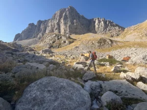 Hiker ascending a rocky alpine trail on Mount Tymfi in the Pindus Mountains, with Gamila Peak and limestone cliffs rising under a clear summer sky.