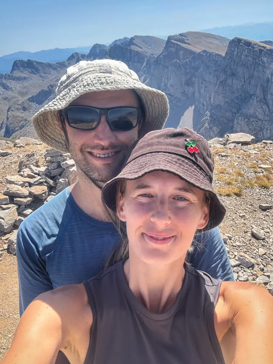 Two hikers taking a selfie on a rocky ridge of Mount Tymfi, with steep limestone cliffs and alpine terrain of the Pindus Mountains in the background.
