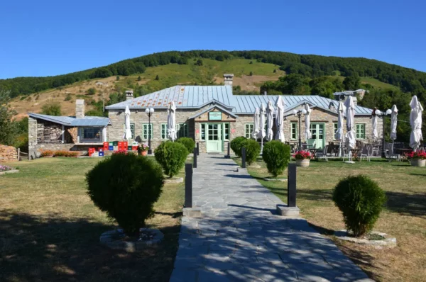 Stone-built hotel in Nymfaio village, Florina, with a central paved walkway, trimmed shrubs, and traditional Macedonian architecture set against forested mountain slopes.