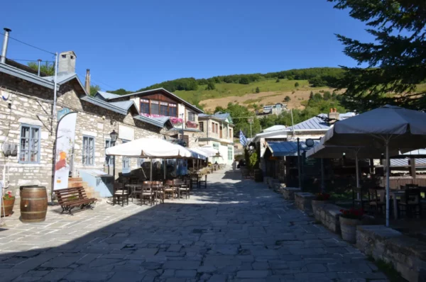 Stone-paved street with cafés and tavern seating in the central square of Nymfaio village, Florina, lined with traditional stone houses under clear mountain light.