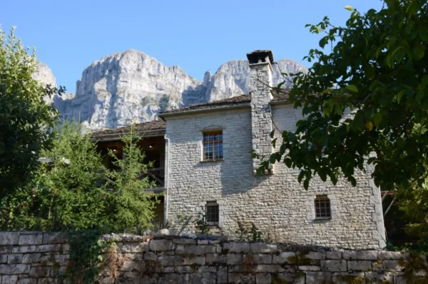 Traditional stone house with a cozy porch surrounded by lush greenery, set against the striking mountain backdrop in Papigo, Zagorochoria, Greece.