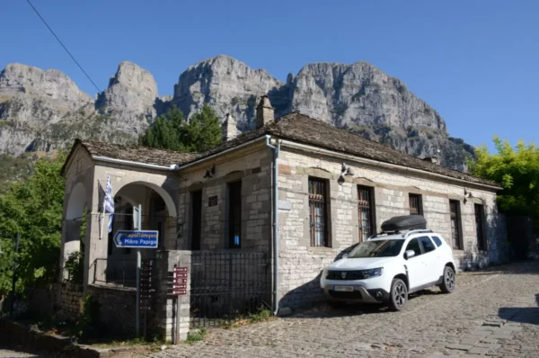 Charming stone building with a sloped roof and a white car parked outside, set against towering mountains in Papigo, Zagorochoria, Greece.