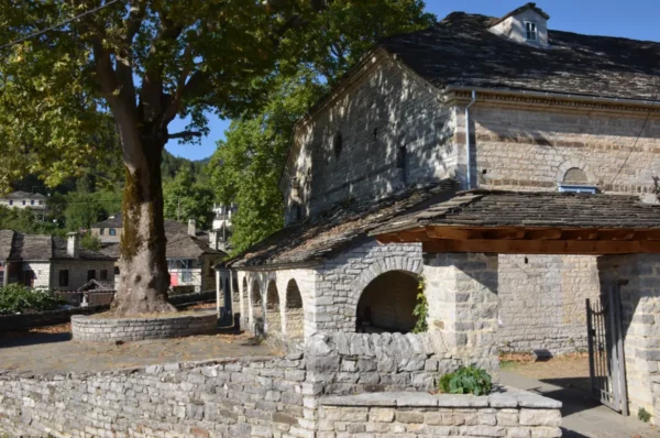 Rustic stone buildings and a large tree create a serene scene in Papigo, Zagorochoria, Ioannina, Greece.