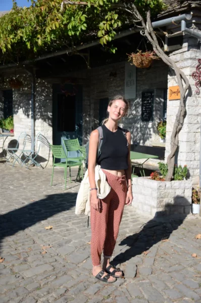 Young woman in casual attire stands in a quaint cobblestone square in Papigo, Zagorochoria, surrounded by greenery and rustic buildings.