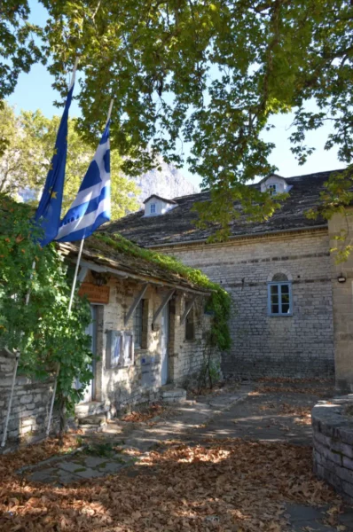 Stone-built school building in Monodendri, Zagorochoria, with a Greek flag in the courtyard, ivy-covered walls, fallen leaves on the stone paving, and trees casting shade.