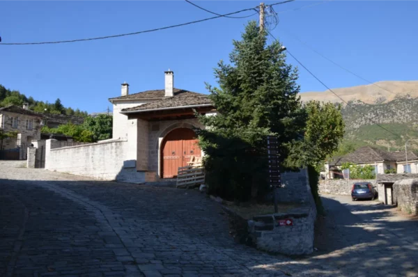 Cobblestone street leading to a charming house with wooden doors and surrounding greenery in Papigo, Zagorochoria, Greece.