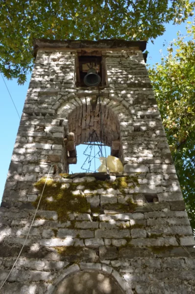 Bell tower with rustic stonework and a clear blue sky surrounded by lush green leaves in Papigo, Zagorochoria, Greece.