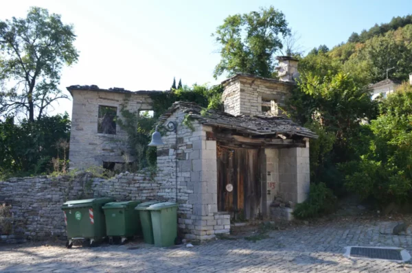 Abandoned stone house with a partially collapsed roof, surrounded by greenery and trash bins in Papigo, Zagorochoria, Greece.