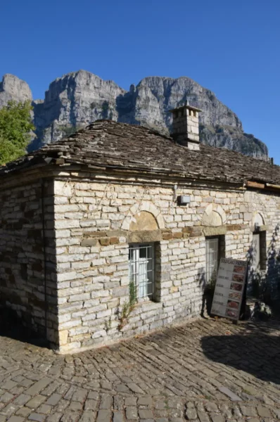Traditional stone building with a sloped roof nestled in the rugged mountains of Zagorochoria, Greece. Clear blue sky above.
