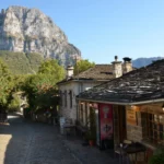 Stone-built houses and cobbled lane in Papigo village, Zagorochoria, Epirus, with limestone cliffs rising behind on a clear summer day.