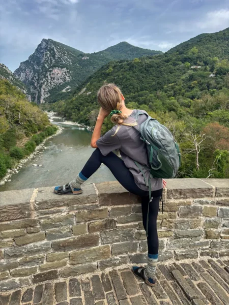 Woman sitting on a stone bridge above the Arachthos River in Tzoumerka, with mountains in the background.