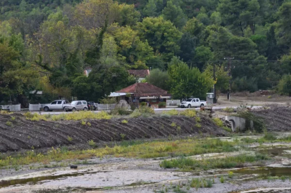 Lush greenery surrounds the small riverside settlement near the Kireas River in Euboea, Greece, with a rustic building and parked cars.