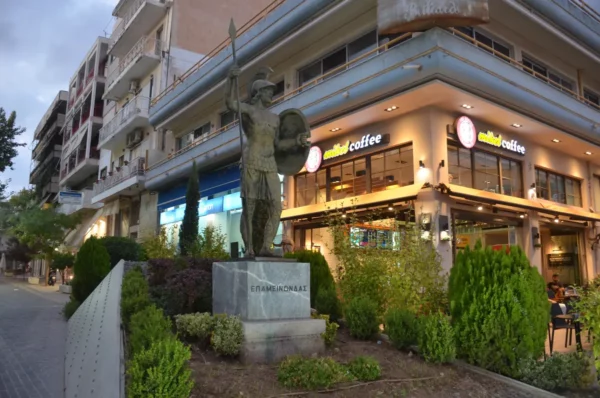 Statue of a soldier stands proudly on a street in Thebes, Boeotia, Greece, surrounded by shops and greenery.