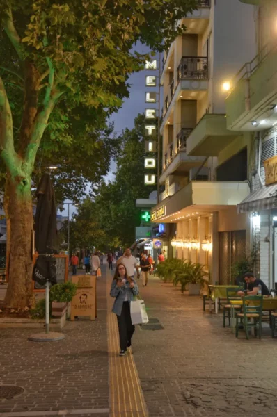 Bustling street scene in Thebes, Boeotia, Greece, featuring shops, greenery, and vibrant evening lights.