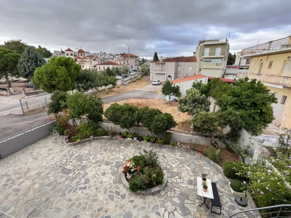Stone courtyard with lush greenery and a view of Thebes, Boeotia, Greece, featuring residential buildings and cloudy skies.